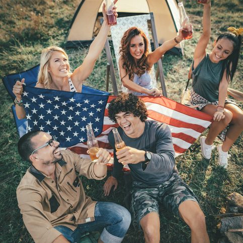 Happy young friends Fourth of July celebration and enjoy a sunny day at the mountain. They're holding american flag, laughing and toasting with beer bottles near tent.
