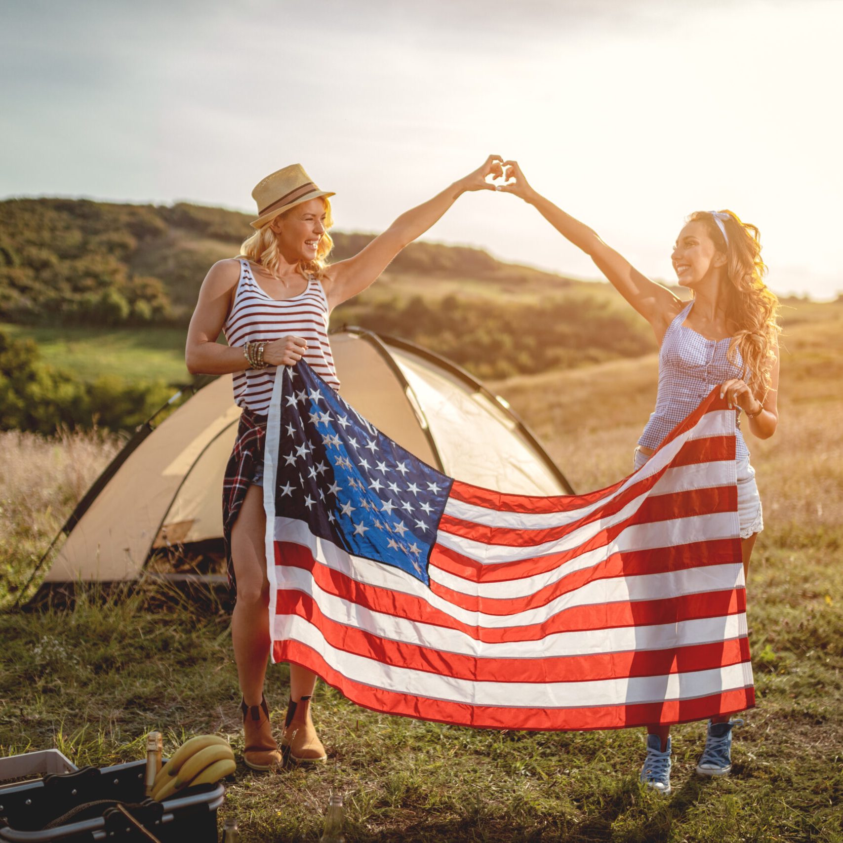 Happy young girl friends enjoys a sunny day in nature. They're holding an american flag in front a campsite tent.