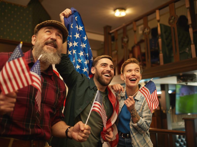 Group of friends with American flags watching football match in sport bar
