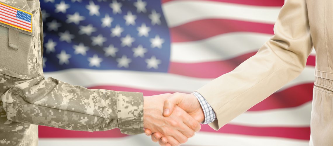 American soldier in uniform and civil man in suit shaking hands with adequate national flag on background - United States of America